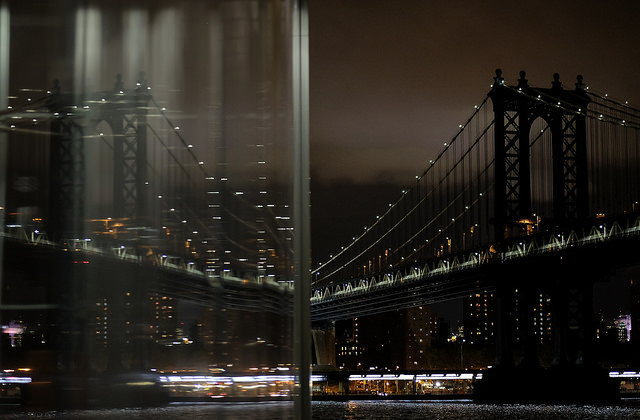 Image of manhattan bridge out of a window with a reflection of the bridge in the other half of the window at night with lights