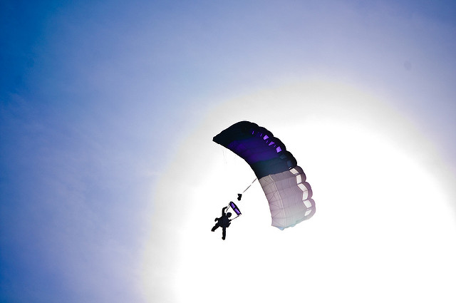 parachute in sky, center of frame with bright sunburst behind