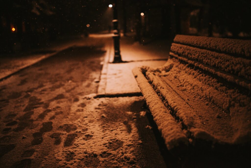 Wooden city bench at dusk covered in snow footprints on sidewalk