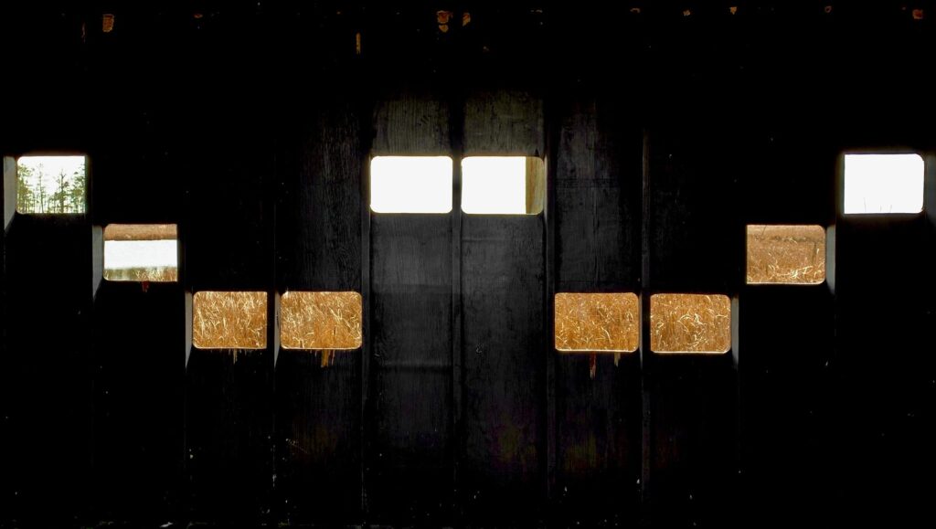 Inside of dark barn with a wall with various windows looking outward hay bales outside
