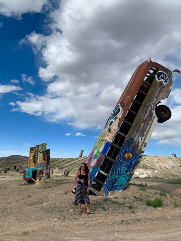 Suzanne Roberts stands in front of a bus that has its nose buried in the sand, and its tail end in the sky.