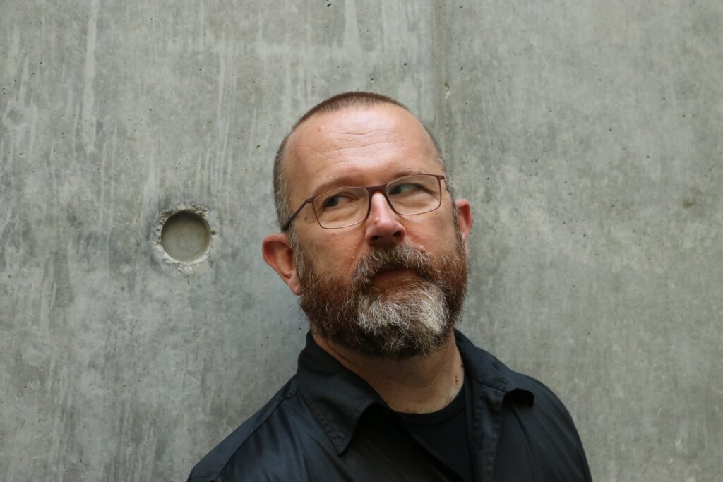 Author Photo: Jonathan Alexander, a white man with glasses and a beard, standing in front of a smooth concrete wall.