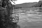 person wearing waders flyfishing in river, focus is on the water with person blurred out