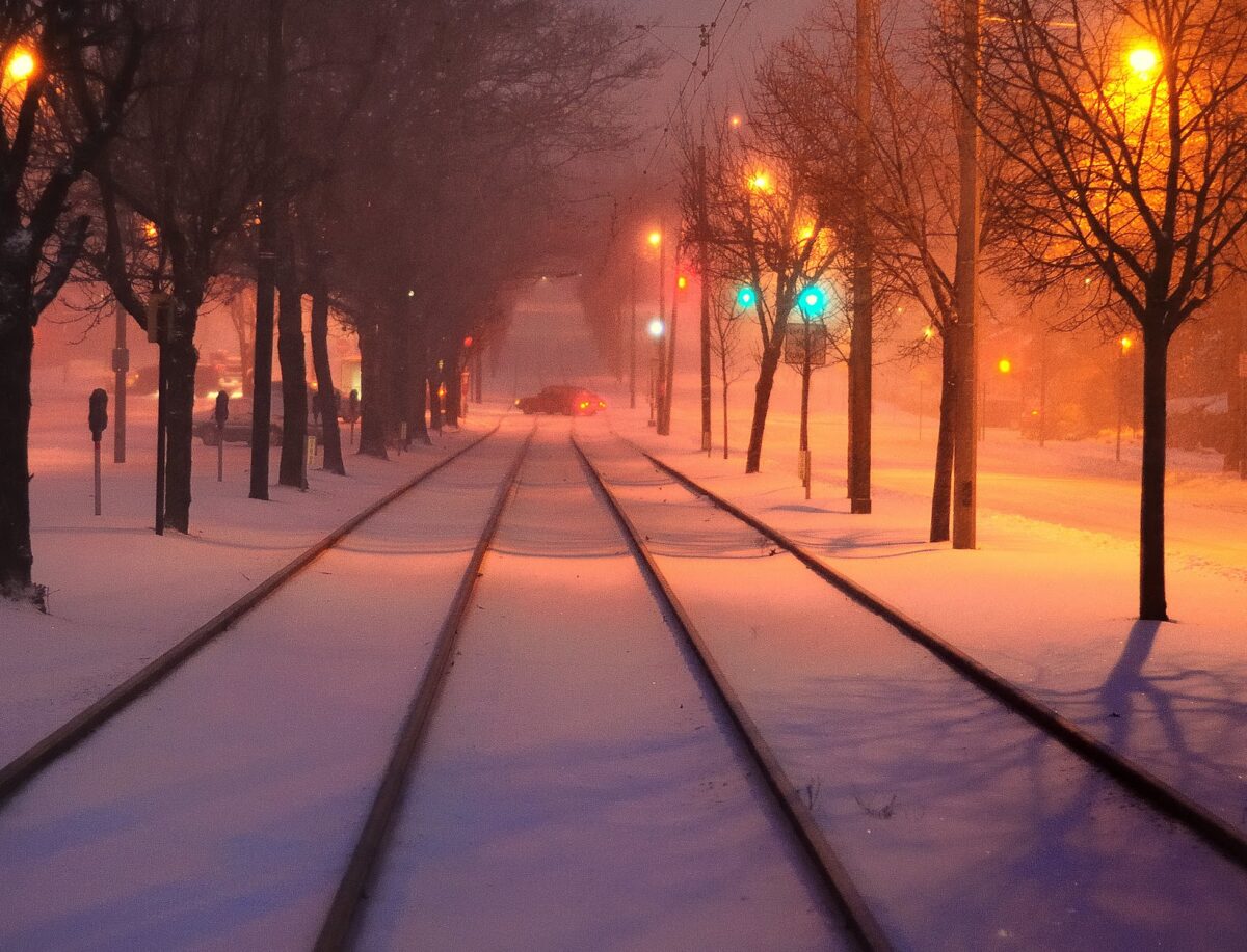 looking down snowy street/rail tracks at dusk; street lights aglow