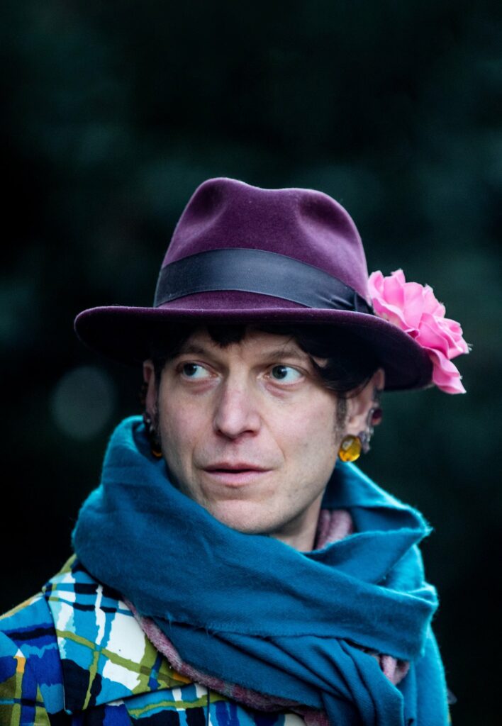 Headshot of Mattilda Bernstein Sycamore, a Jewish woman in purple hat with pink flower, blue scarf.