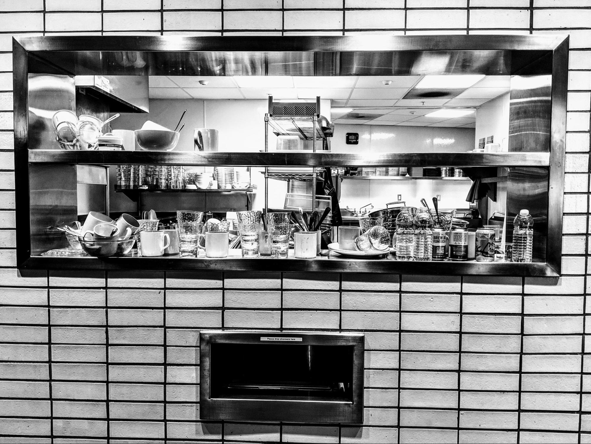 looking through window and counter of a cafeteria kitchen with bussed plates, cups and items in view; modern, sterile kitchen design