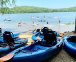 busy day at the lake with lots of boats and kayaks in the water and on the beach. Green hills in the distance