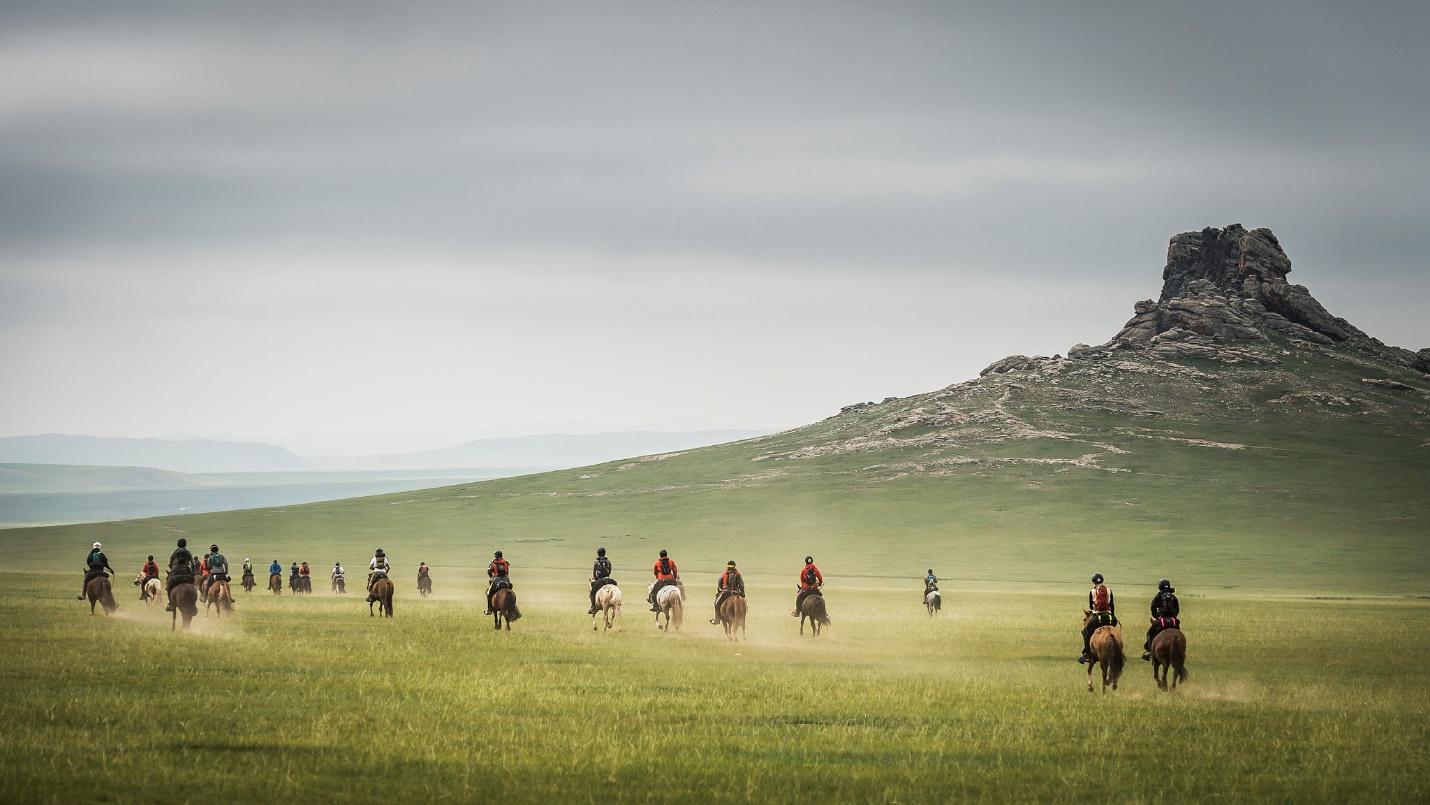 A group of people riding horses in a field, with a butte in the background